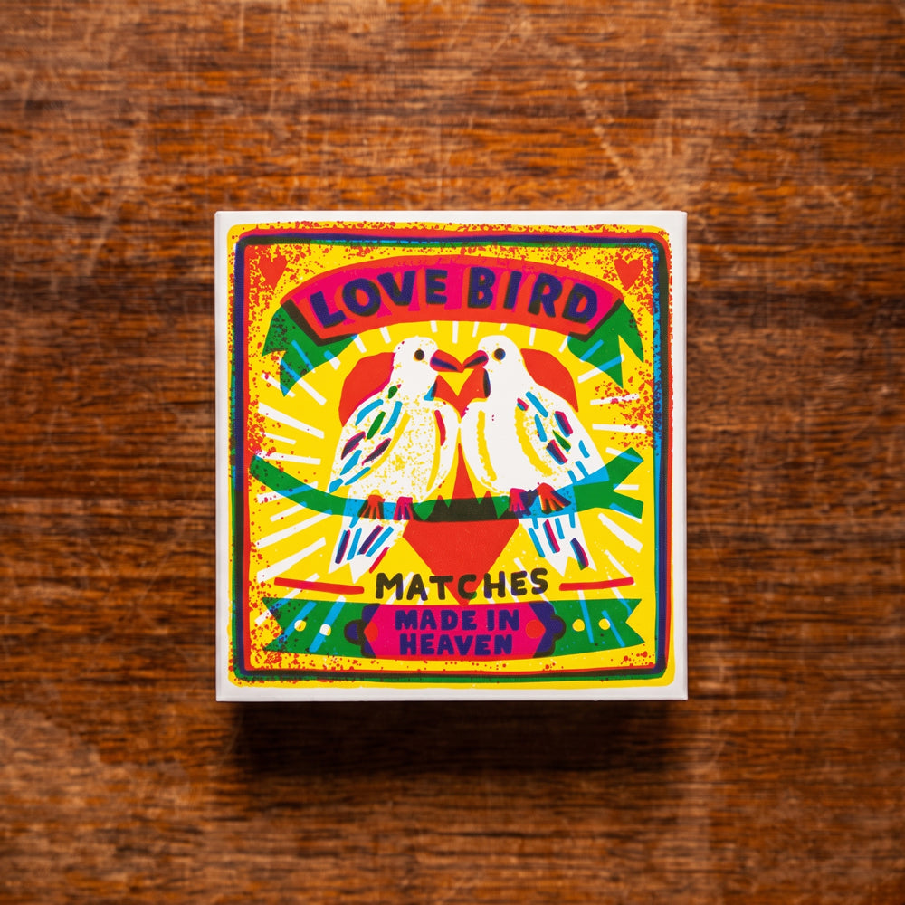 A square matchbox with a colorful vintage-style illustration of two doves and the text 'LOVE BIRD MATCHES MADE IN HEAVEN,' placed on a brown wooden surface.