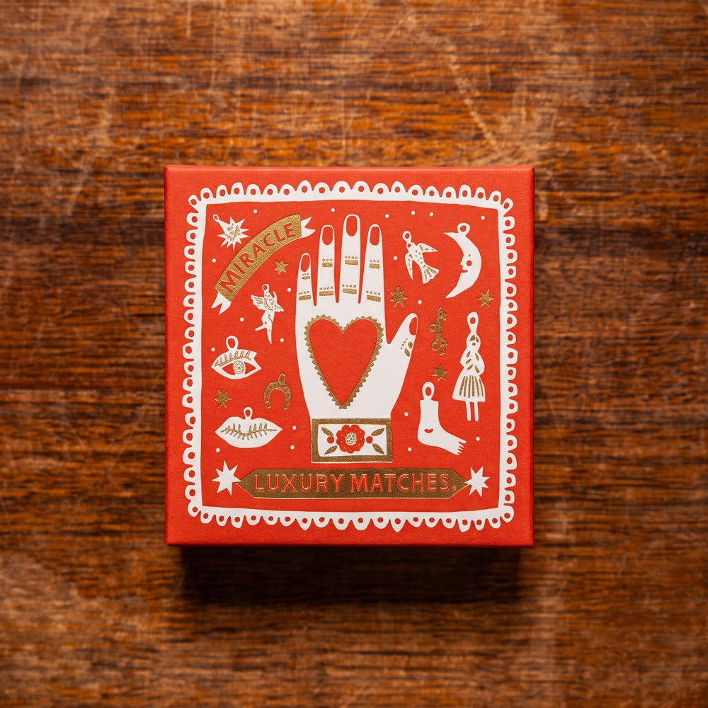 A square red box of 'Miracle Luxury Matches' with a white hamsa hand illustration and a red heart in the center, placed on a rustic wooden surface.