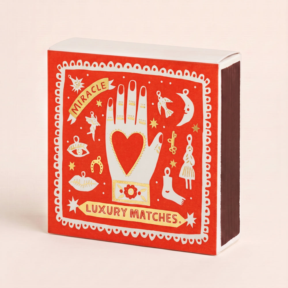 A red matchbox-style box of luxury matches with white and gold illustrations, including a hand with a heart, stars, and decorative borders. The front reads 'Miracle Luxury Matches' and the side shows dark brown match heads.