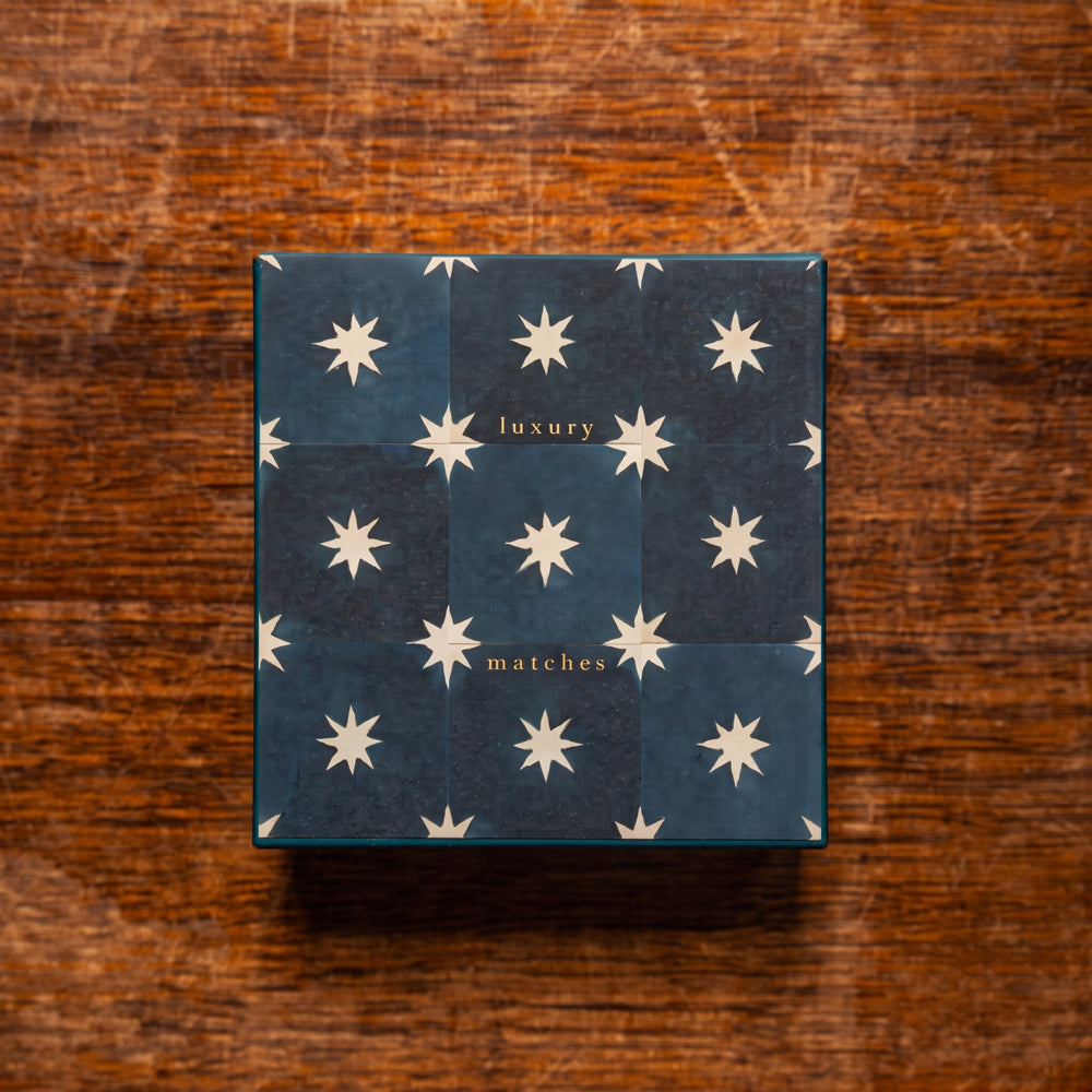 A square box of luxury matches with a dark navy blue cover decorated with a grid of cream-colored starburst patterns and gold lettering, placed on a rustic brown wooden surface.