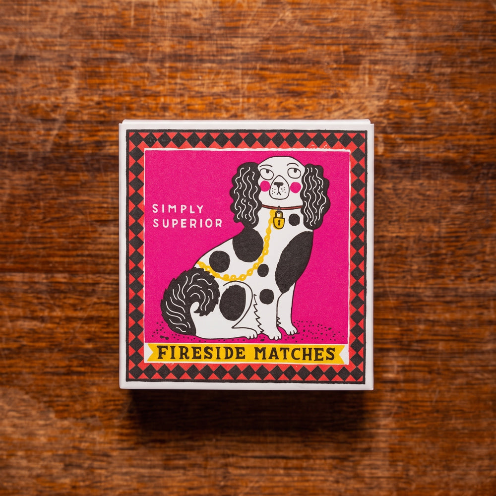 A square box of Fireside Matches featuring a stylized black and white spotted dog illustration on a bright pink background, placed on a brown wooden surface.