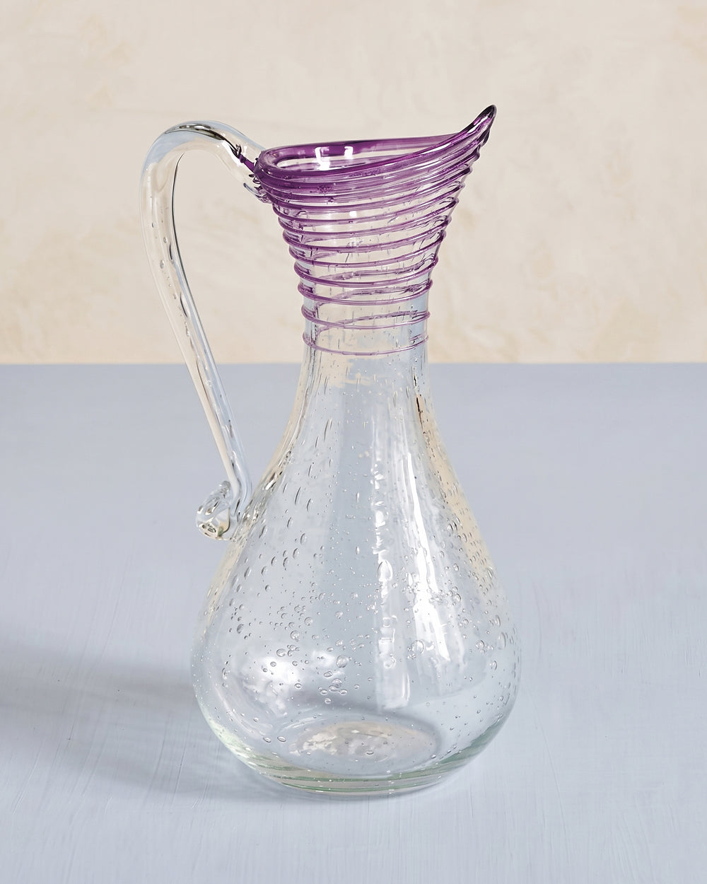 Vintage clear bubble glass pitcher in Biot style with a purple ribbed spout, shown on a light grey background, available at Wildschut Antiques.