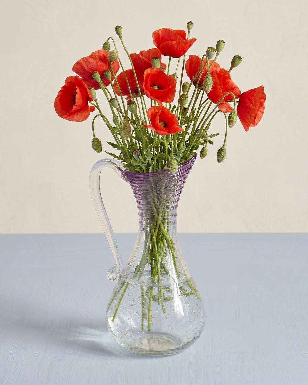 Vintage clear bubble glass pitcher in Biot style with a purple ribbed spout, shown with a bouquet of red poppies, available at Wildschut Antiques.
