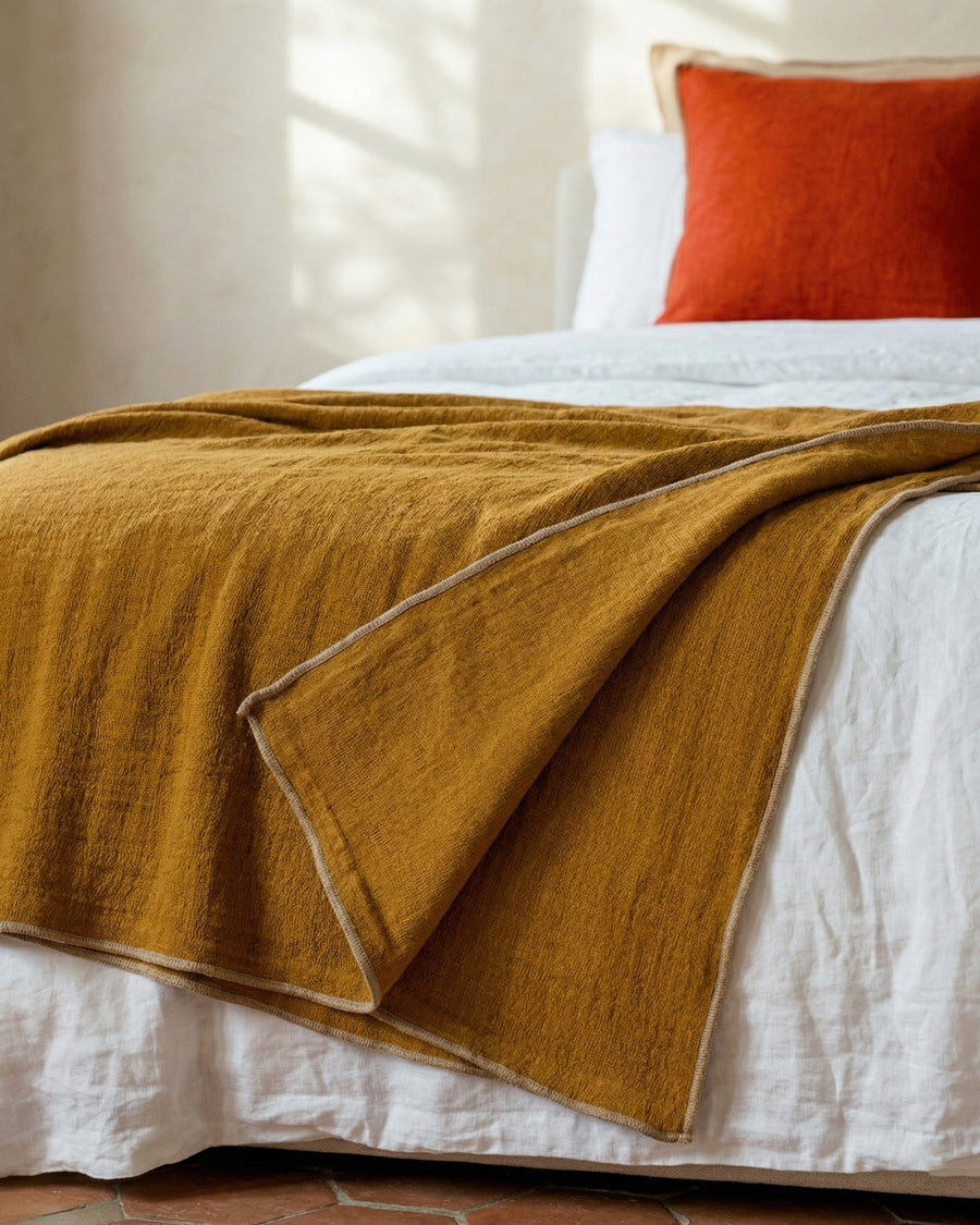 Ginger brown blanket draped over a bed with white sheets and a red pillow.