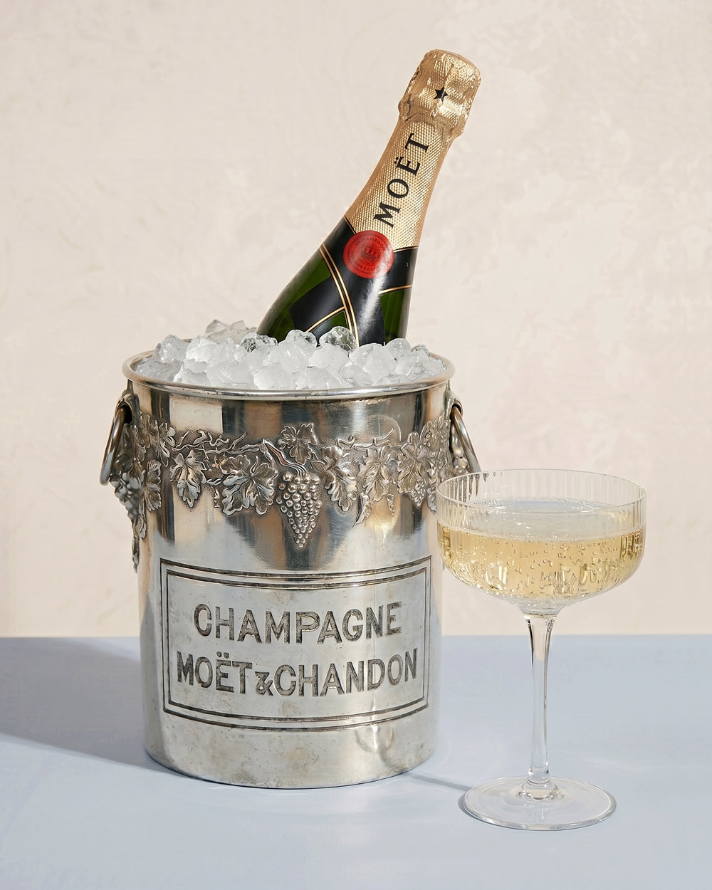 Moét & Chandon champagne bottle in a vintage silver plated ice bucket with Moét & Chandon branding and a glass of champagne on a light background. Available at Wildschut Antiques.