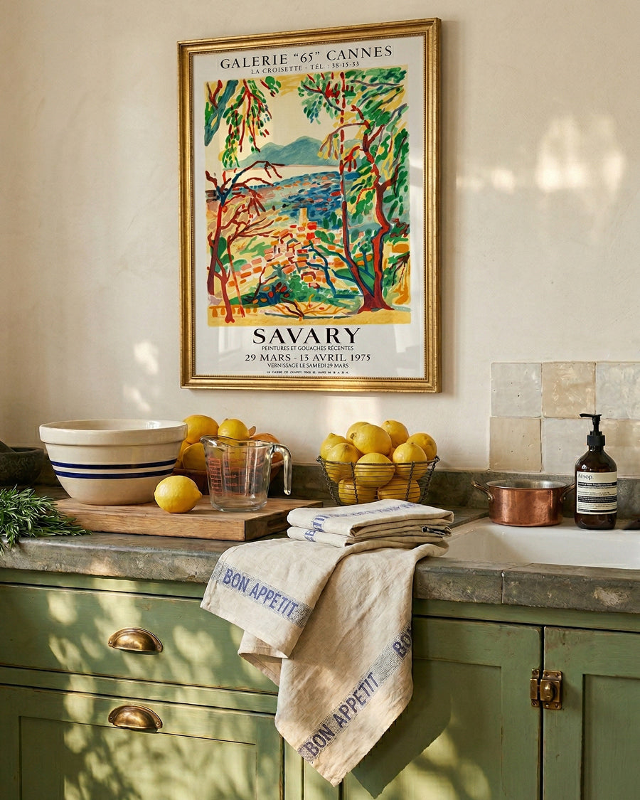 Kitchen counter with lemons, linen 'Bon Appetit' towels in blue, and a colorful poster on the wall by Savary. Linen towels by Charvet Editions available via Wildschut Antiques.