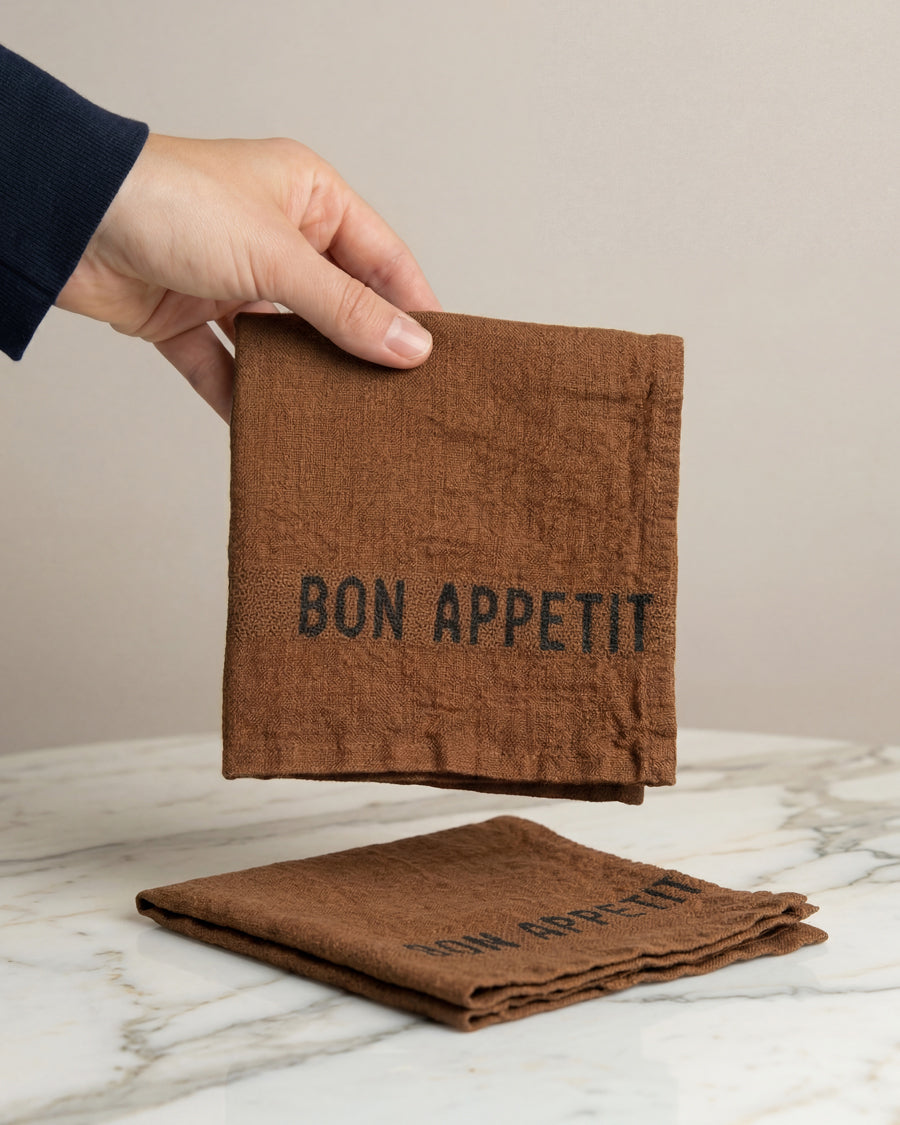Brown linen napkin with 'Bon Appetit' text held by a hand on a marble surface, by Charvet Editions available via Wildschut Antiques.