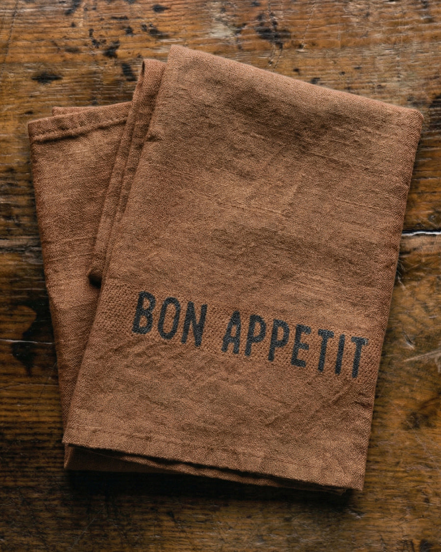 Brown linen napkin with 'Bon Appetit' text on a wooden surface, by Charvet Editions available via Wildschut Antiques.