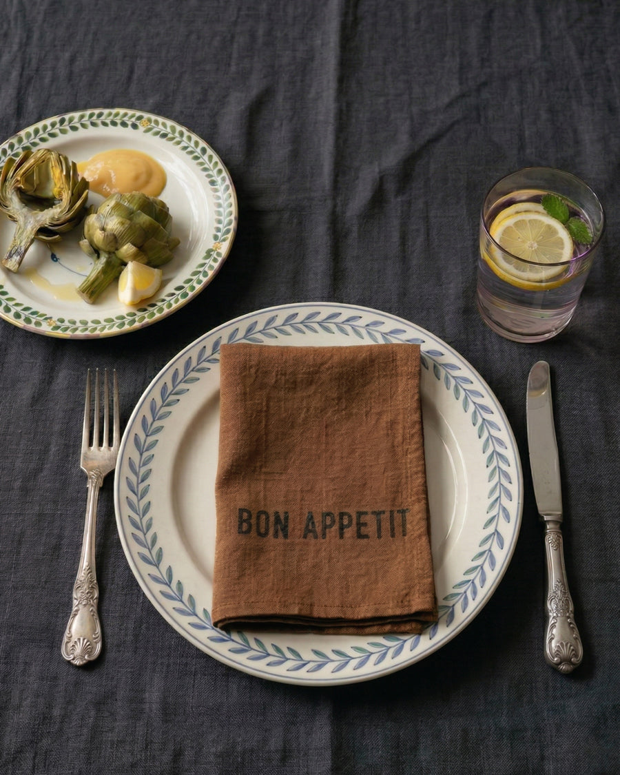 Dining table setting with a brown 100% linen 'Bon Appetit' napkin, plate, fork, knife, and glass of water. Napkin by Charvet Editions available via Wildschut Antiques.