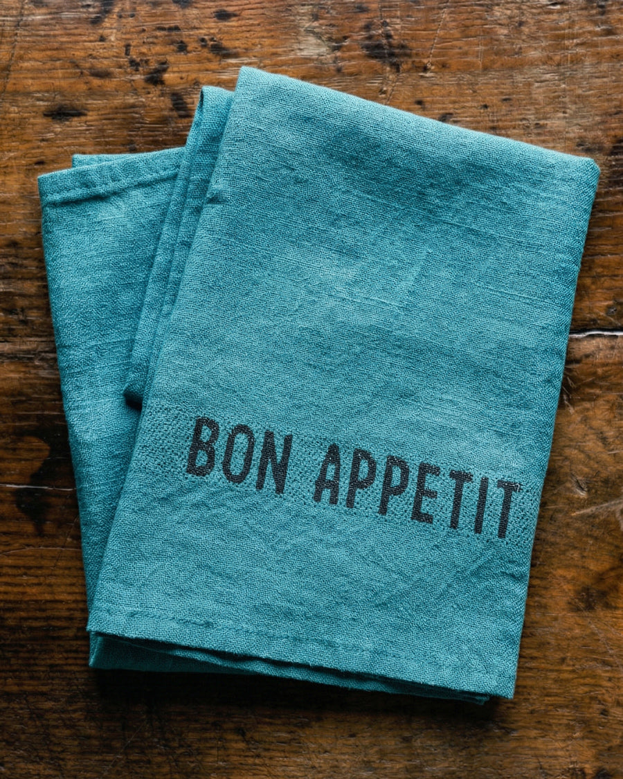 Teal linen napkin with 'Bon Appetit' text on a wooden surface, by Charvet Editions available via Wildschut Antiques.