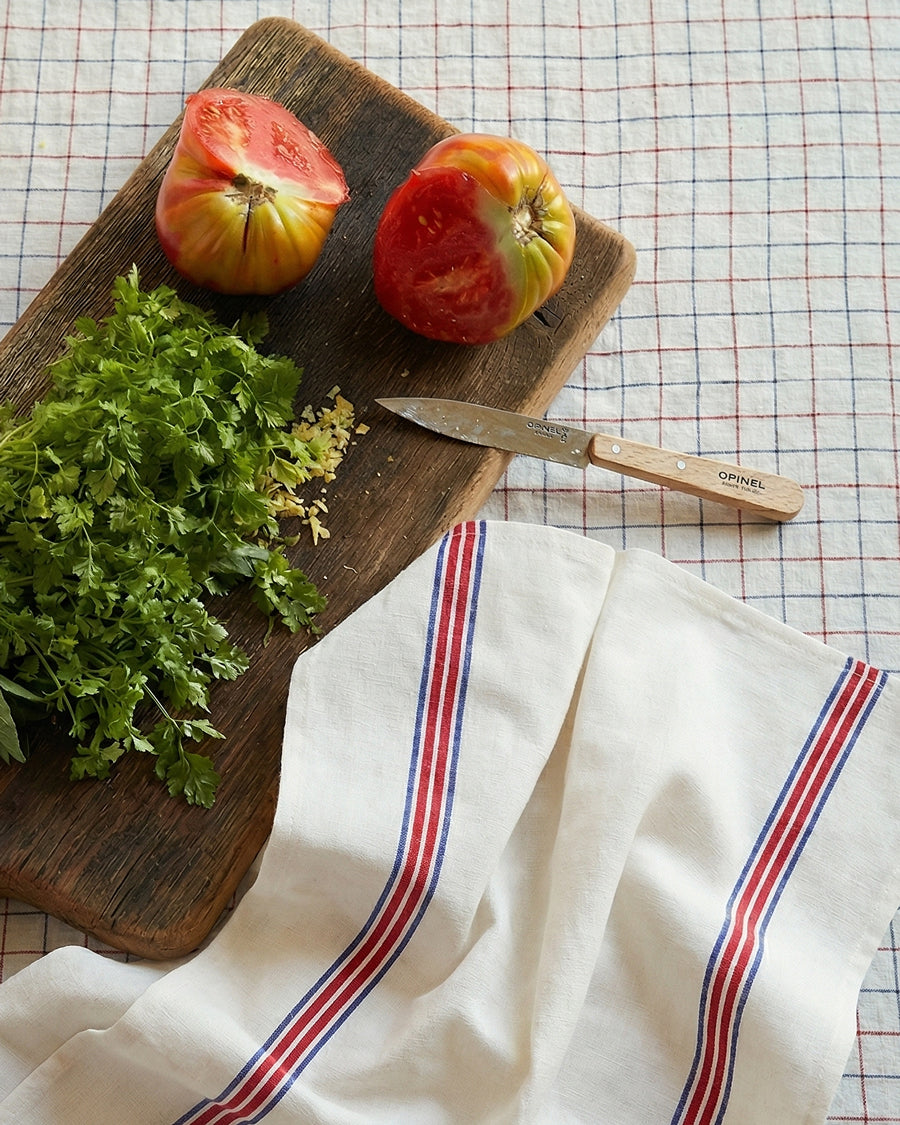 Wooden cutting board with tomatoes, herbs, and a knife on a checkered tablecloth, next to a towel with blue and red stripes by Charvet Editions, available via Wildschut Antiques. 