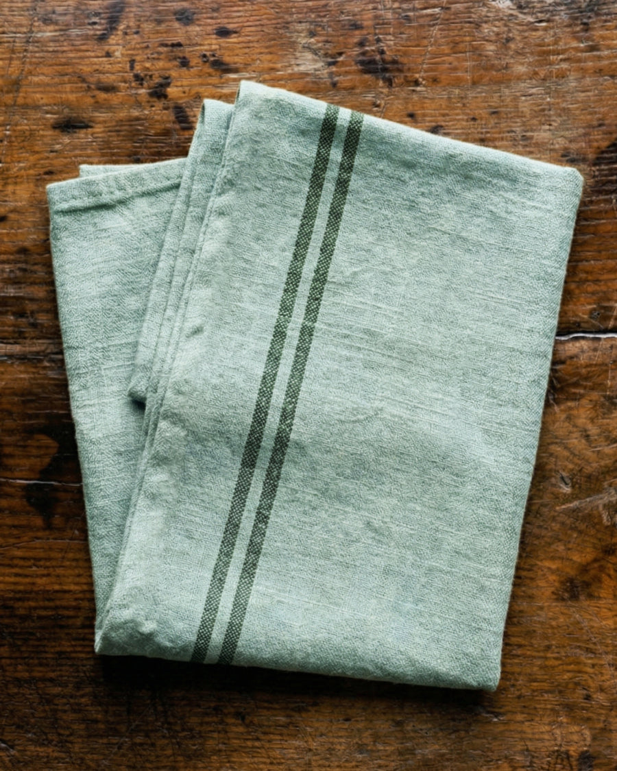 Folded mint green towel with dark stripes on a wooden surface. Linen country towel by Charvet Editions available via Wildschut Antiques.