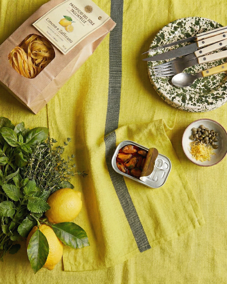 Table setting with a can of food, lemons, herbs, and pasta on a yellow tablecloth and lemon yellow linen towel with a dark stripe. Linen towel by Charvet Editions available via Wildschut Antiques.