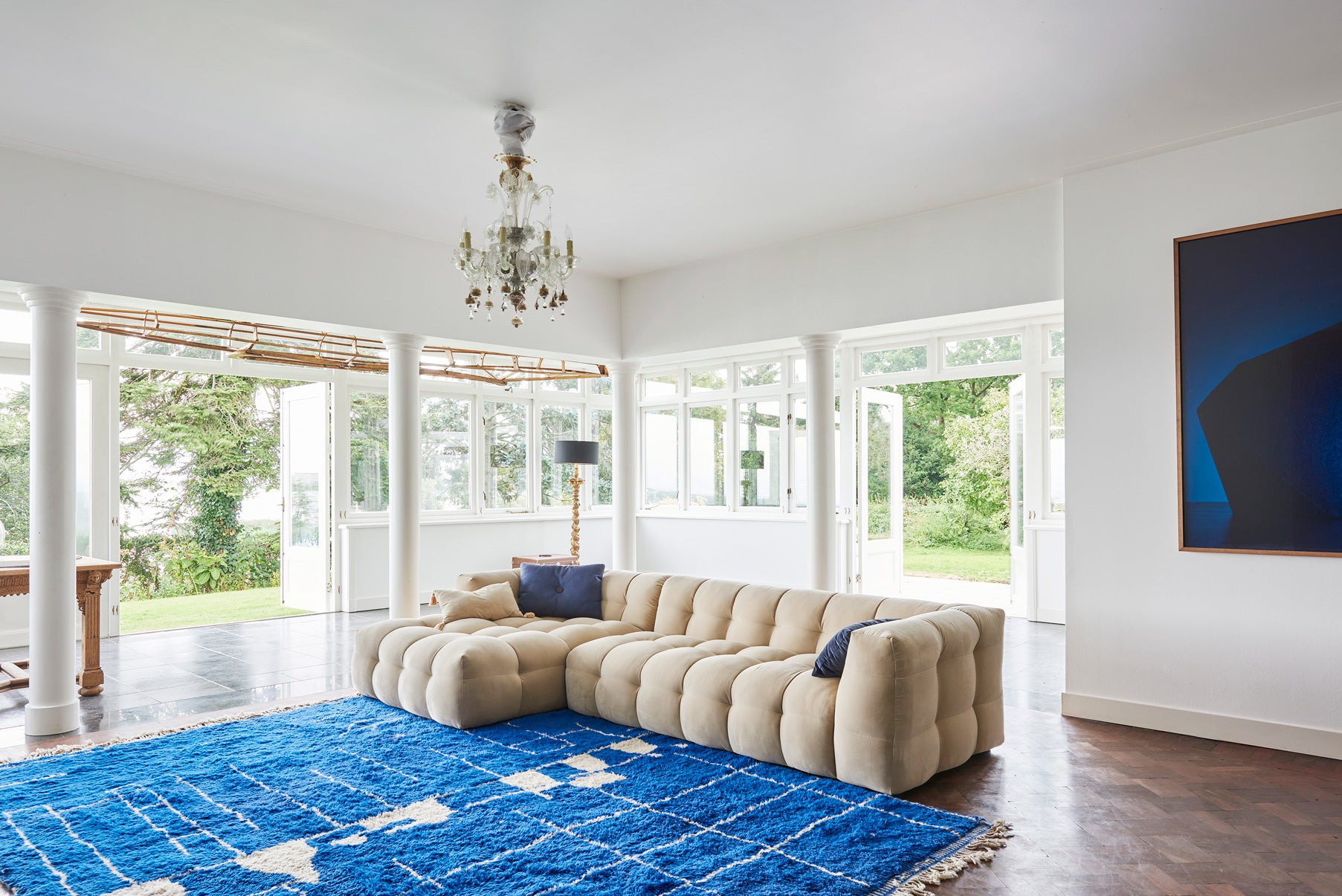 Spacious, light-filled room with a dark wooden floor and a soft cream-colored sofa. A large vintage cobalt blue Moroccan rug with asymmetrical white lines anchors the space. A Venetian chandelier hangs from the ceiling, while a cobalt blue photograph adds a bold touch to the wall.