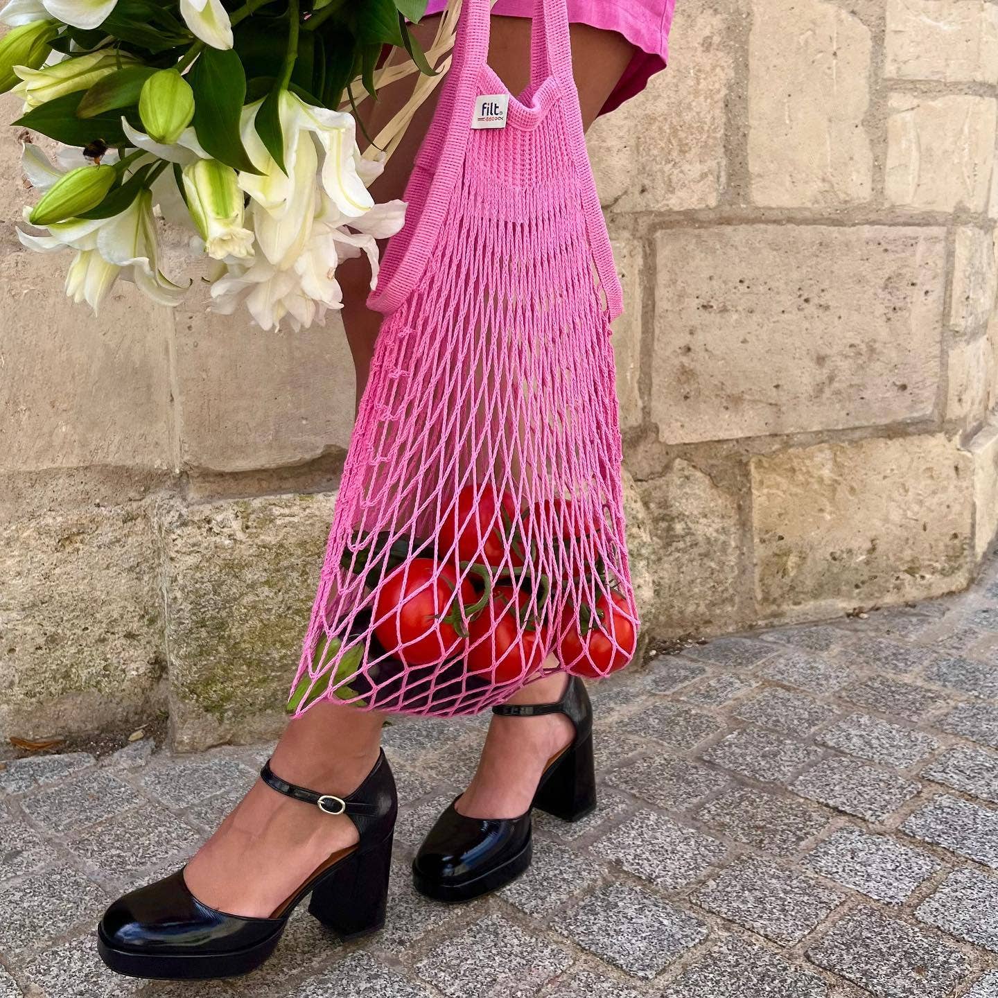 Person holding a pink mesh bag with tomatoes and flowers against a stone wall.