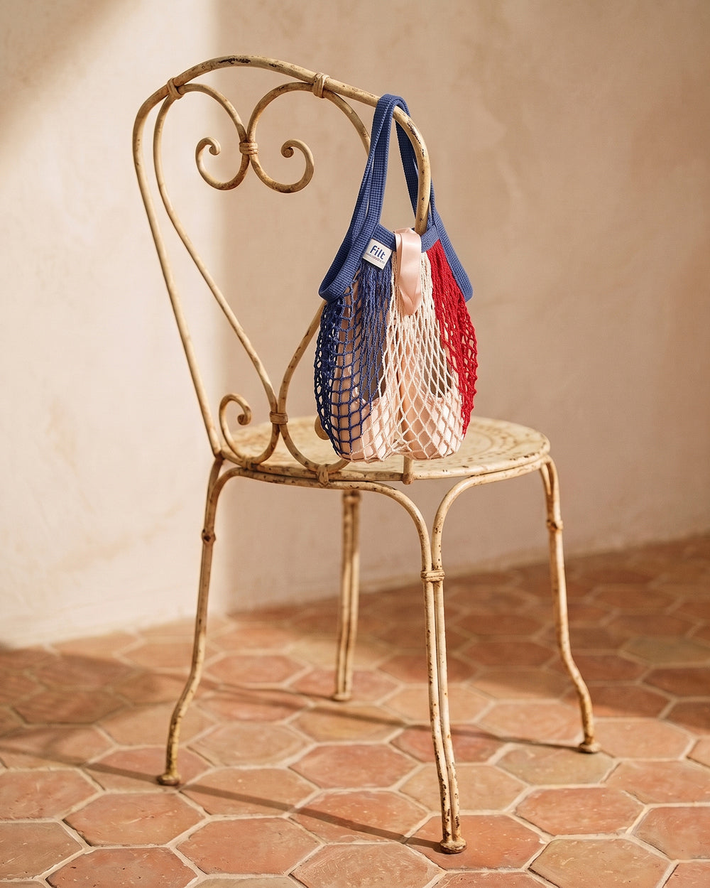 A red, white, and blue reusable mesh tote bag with shoulder straps hangs from a distressed gold metal chair in a warm, sunlit room with terracotta tile flooring. By Filt 1860, available at Wildschut Antiques & Decor.