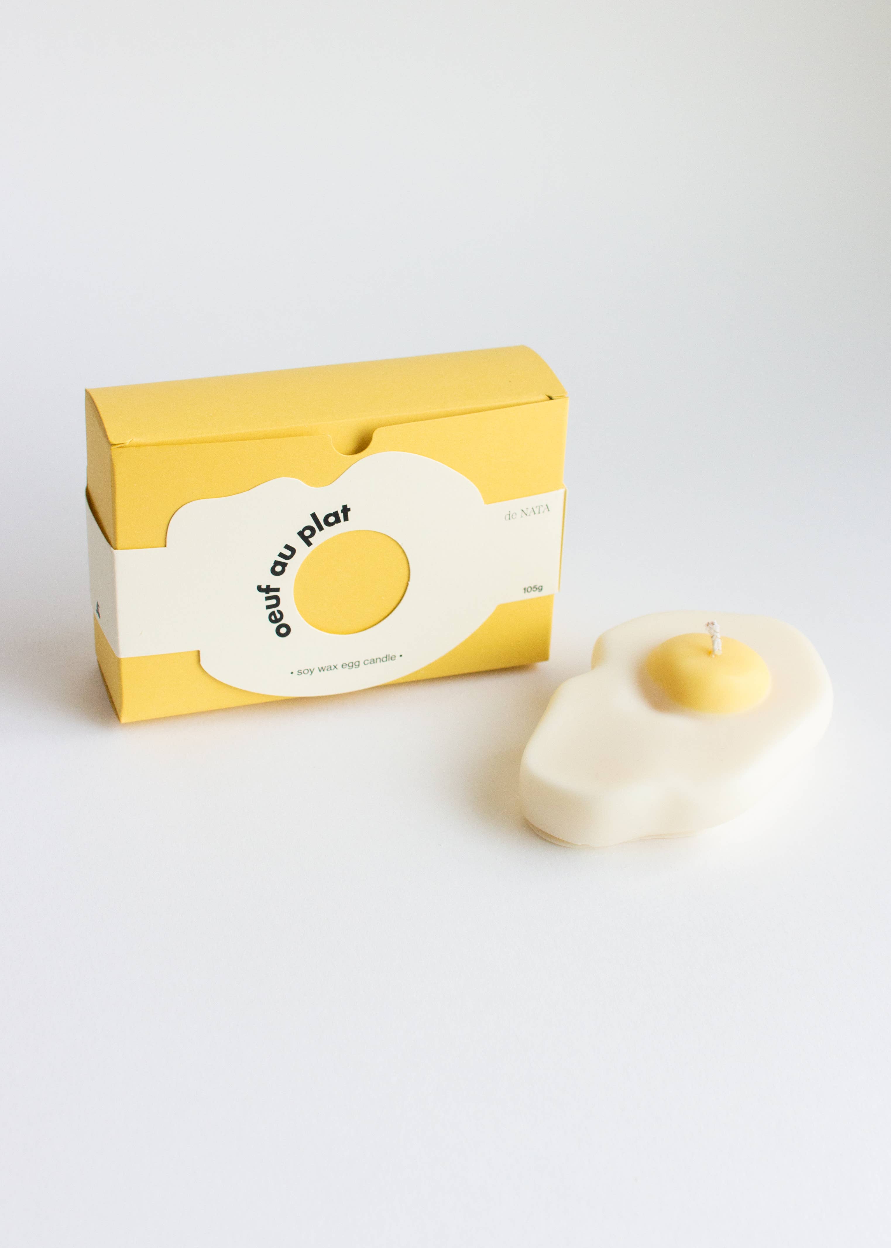 Fried egg-shaped candle with a yellow center next to its packaging on a white background. Part of the Playful Food Candle Collection available via Wildschut Antiques Concept Store 