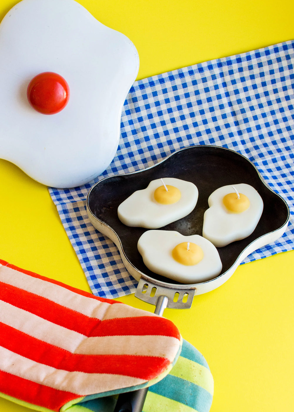 Fried egg-shaped candles in a mini frying pan on a checkered tablecloth with a yellow background.