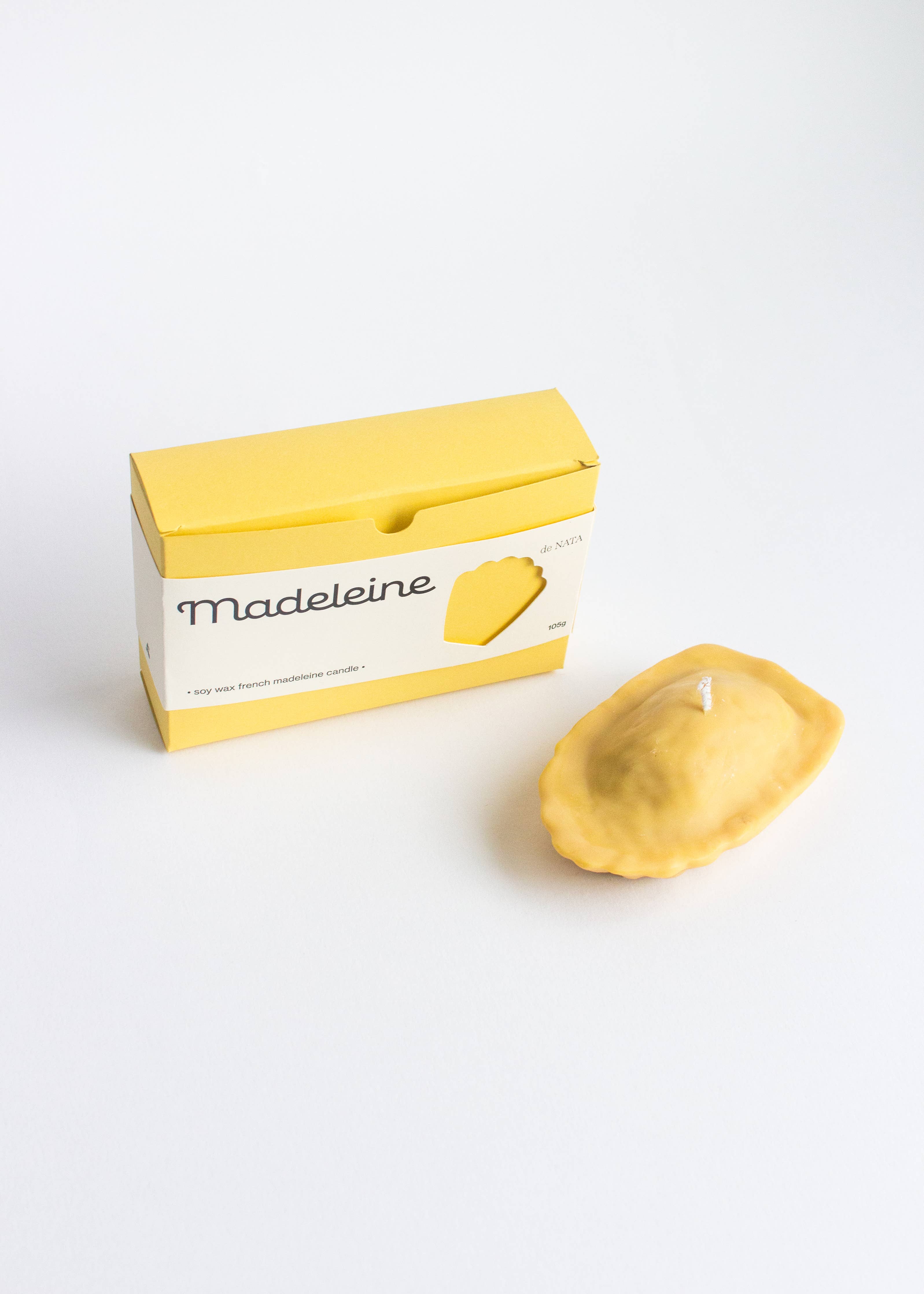 Yellow packaging labeled 'Madeleine' next to a candle in the shape of a Madeleine cake on a white background.