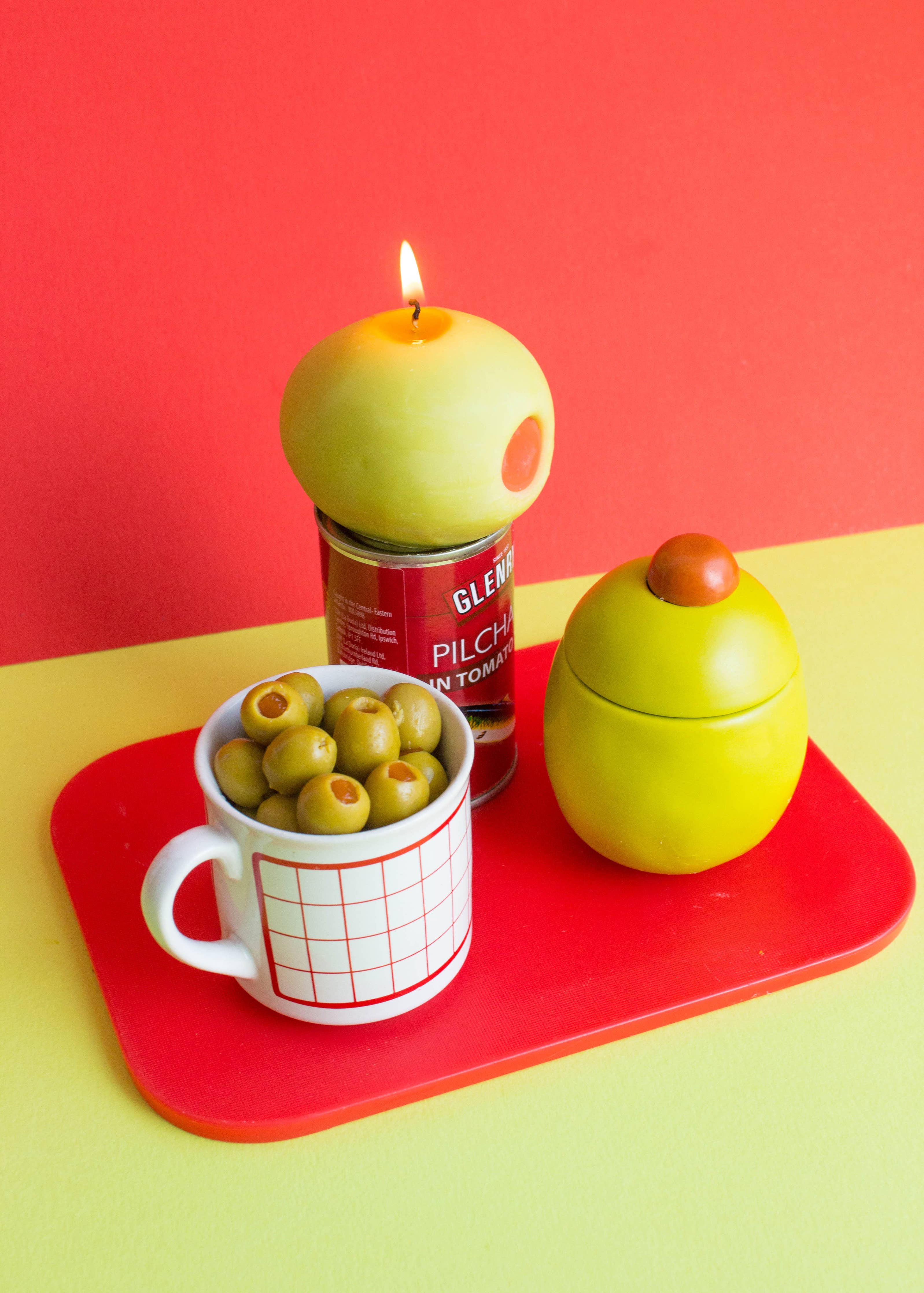Candle shaped like a green olive, on top of a red can, and a cup with olives on a red tray against a red background.