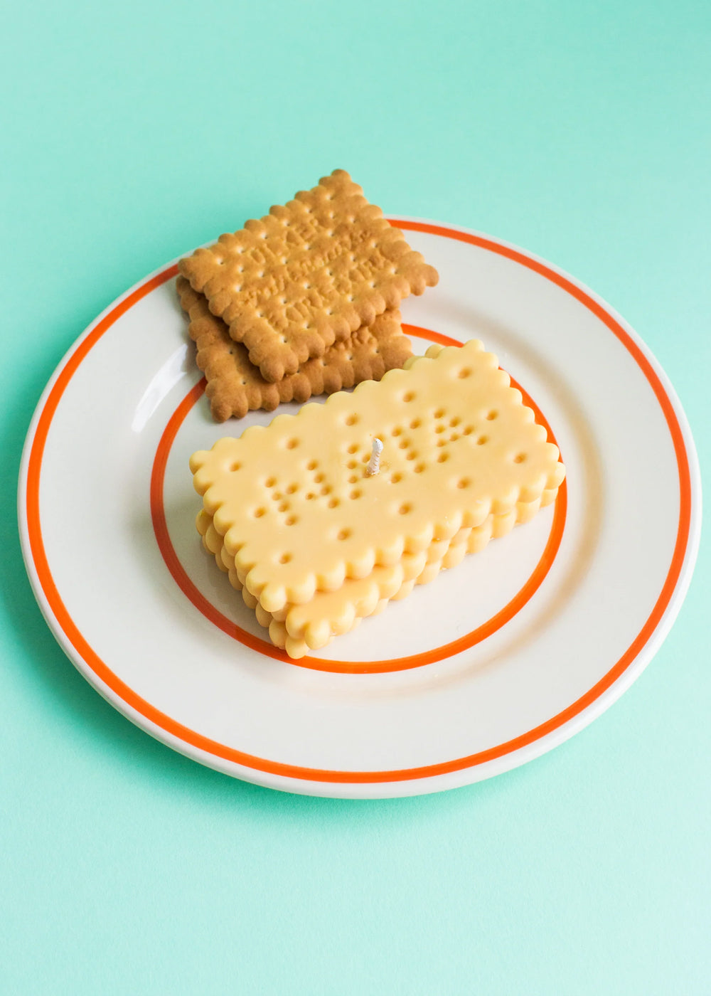 Two biscuits next to a candle in the shape of stacked biscuits on a white plate with an orange rim against a mint green background