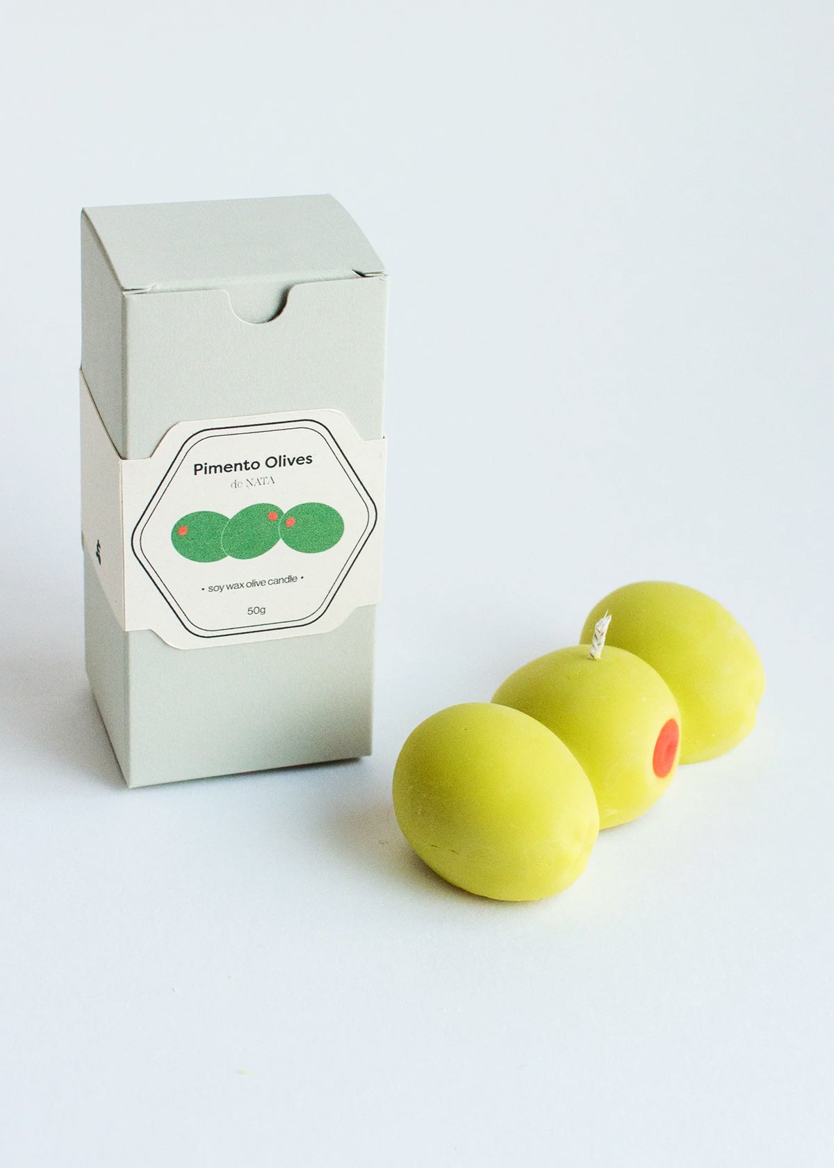 A light green candle in the shape of three  olives with a red center next to a box labeled 'Pimento Olives' on a white background. Part of the Playful Food Candle Collection available via Wildschut Antiques Concept Store