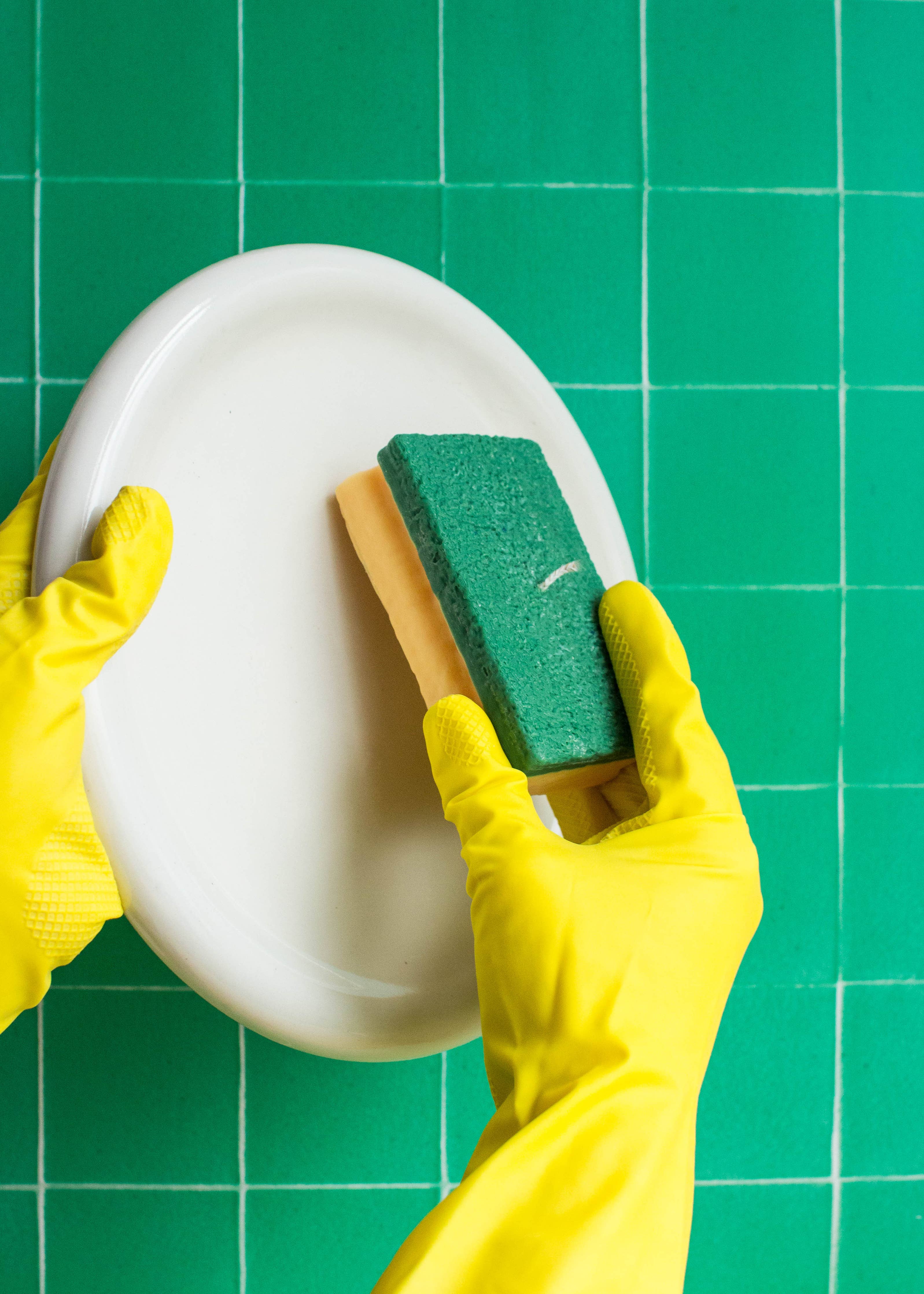 Person cleaning a white plate with a green sponge on a green grid background. Fun fact: the sponge is not real, it is a candle!
