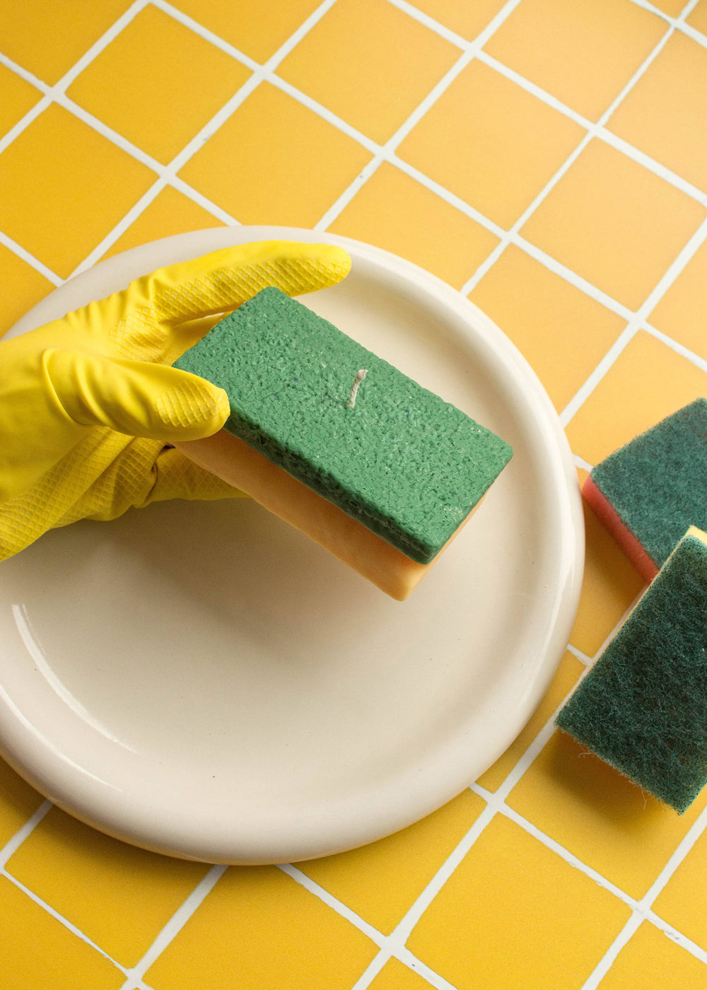 Hand wearing a yellow glove holding a green sponge over a white plate on a yellow tiled floor. Fun fact: the sponge is not real, it is a candle in the shape of a sponge!