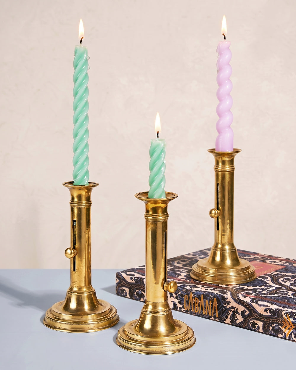 Three antique French brass candlesticks with pastel twisted candles on a neutral background. Available at Wildschut Antiques.