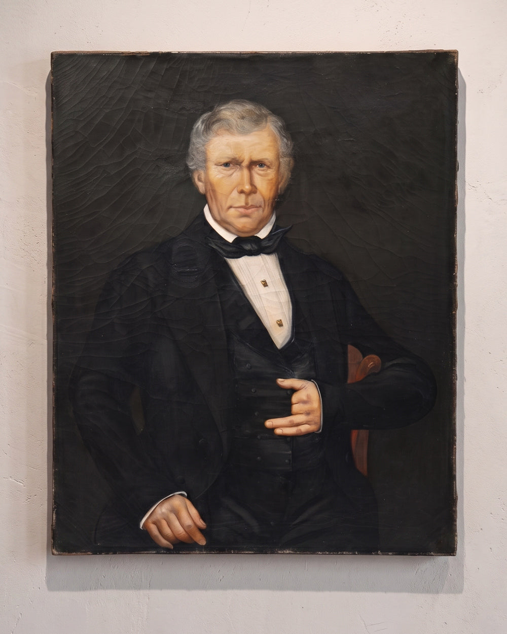 An antique portrait oil painting of an elderly man in a black formal suit and bow tie against a dark background, mounted on a light wall. Available via Wildschut Antiques.