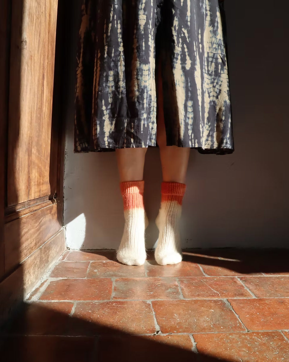 Person wearing a patterned skirt, white wool socks with orange accents, on a red brick floor.