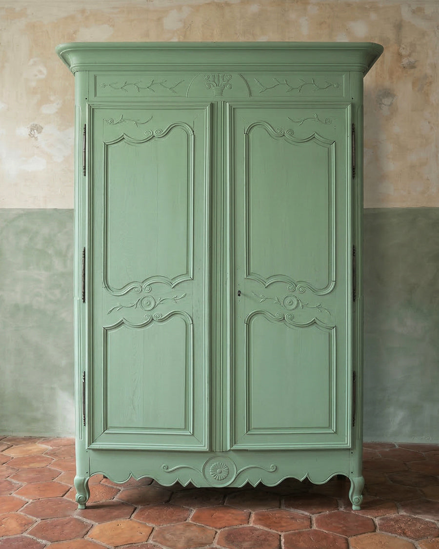 Antique French green ornate cabinet or marriage armoire against a textured wall with a terracotta tiled floor, available via Wildschut Antiques.