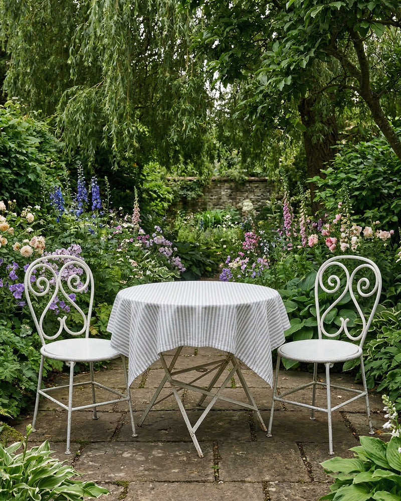 Vintage French white garden table and chairs with a striped tablecloth in a lush garden setting, available via Wildschut Antiques.
