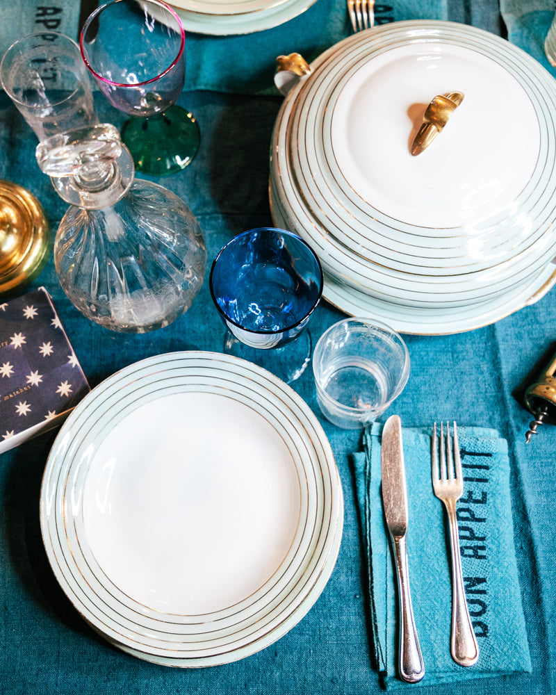 Dining table setting with white plates, silverware, and a blue napkin on a teal tablecloth.