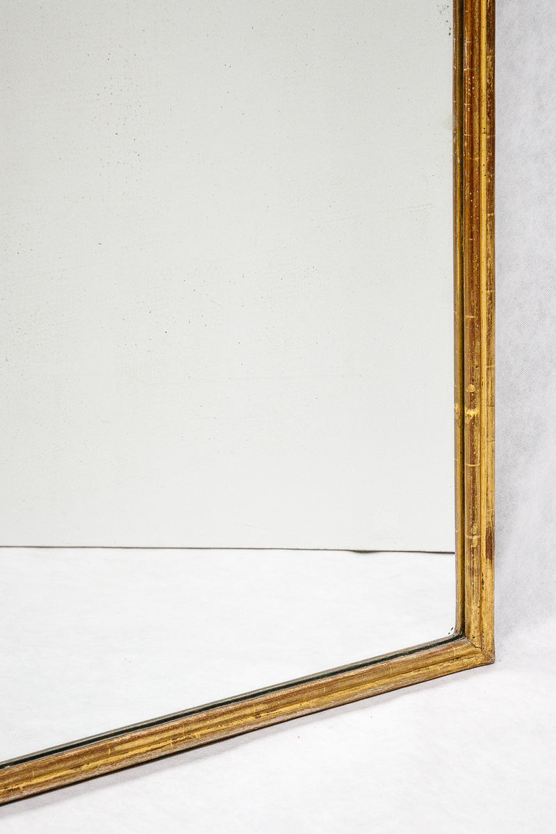 Close-up of a bottom corner of a rectangular antique French mirror with a slender gold frame, set against a white background