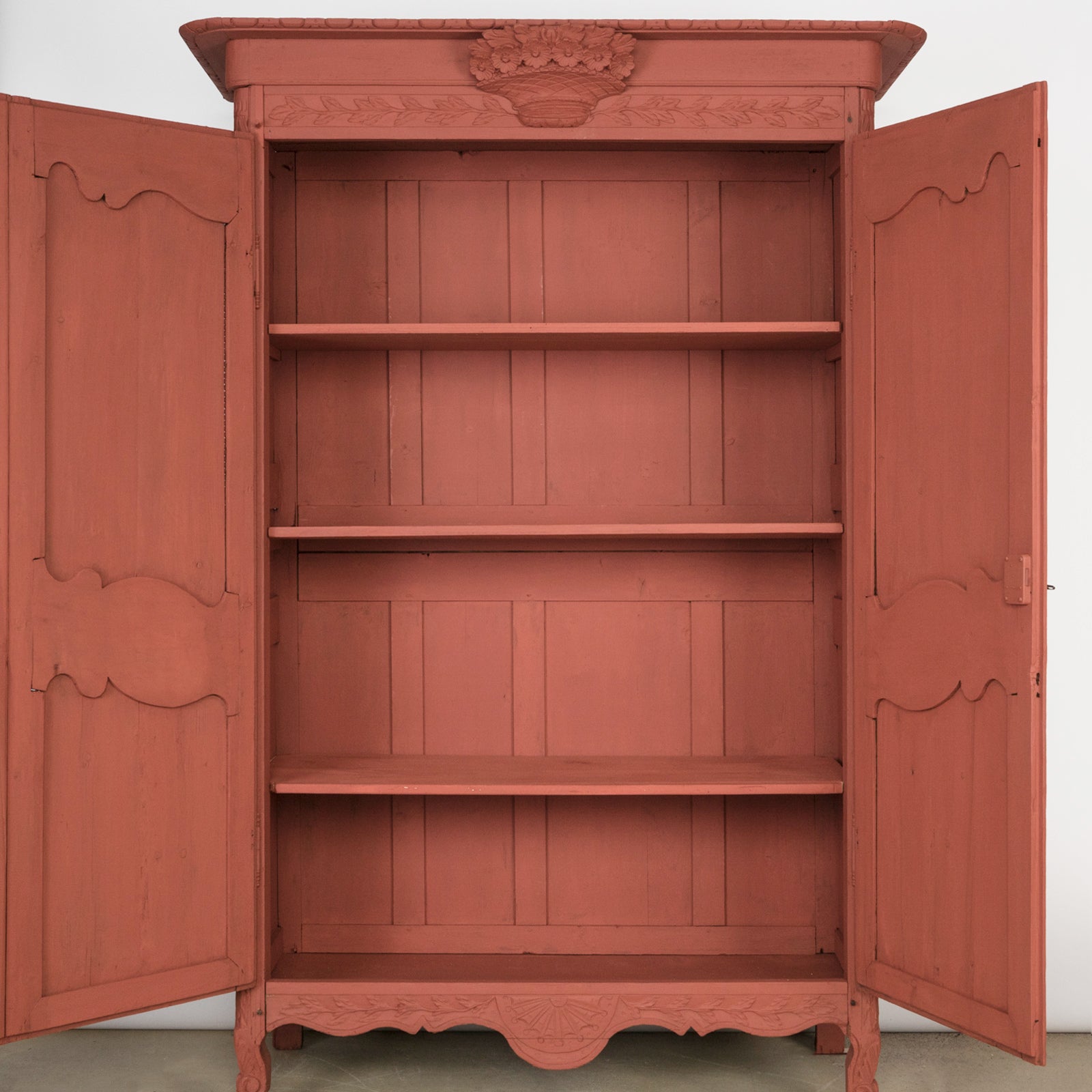 Interior of an antique French marriage armoire, painted in vibrant brick red, displayed with two open doors revealing three shelves.