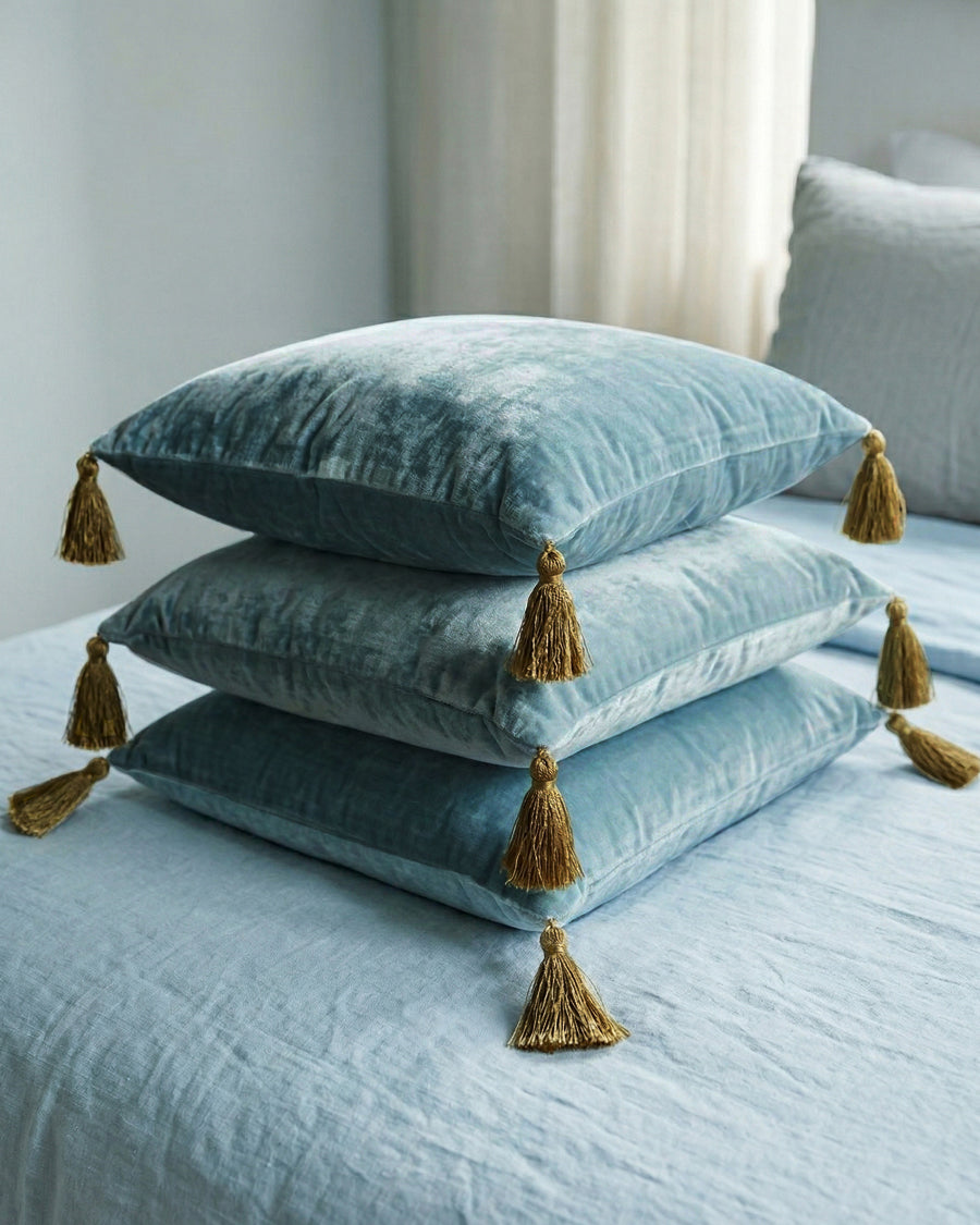 Stack of light grey-blue velvet pillows with olive green tassels on a bed, by Monde Sauvage available via Wildschut Antiques.