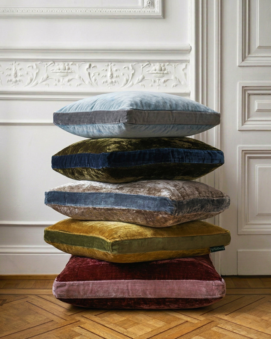 Stack of colorful velvet cushions on a wooden floor with decorative wall in the background, by Monde Sauvage available via Wildschut Antiques.