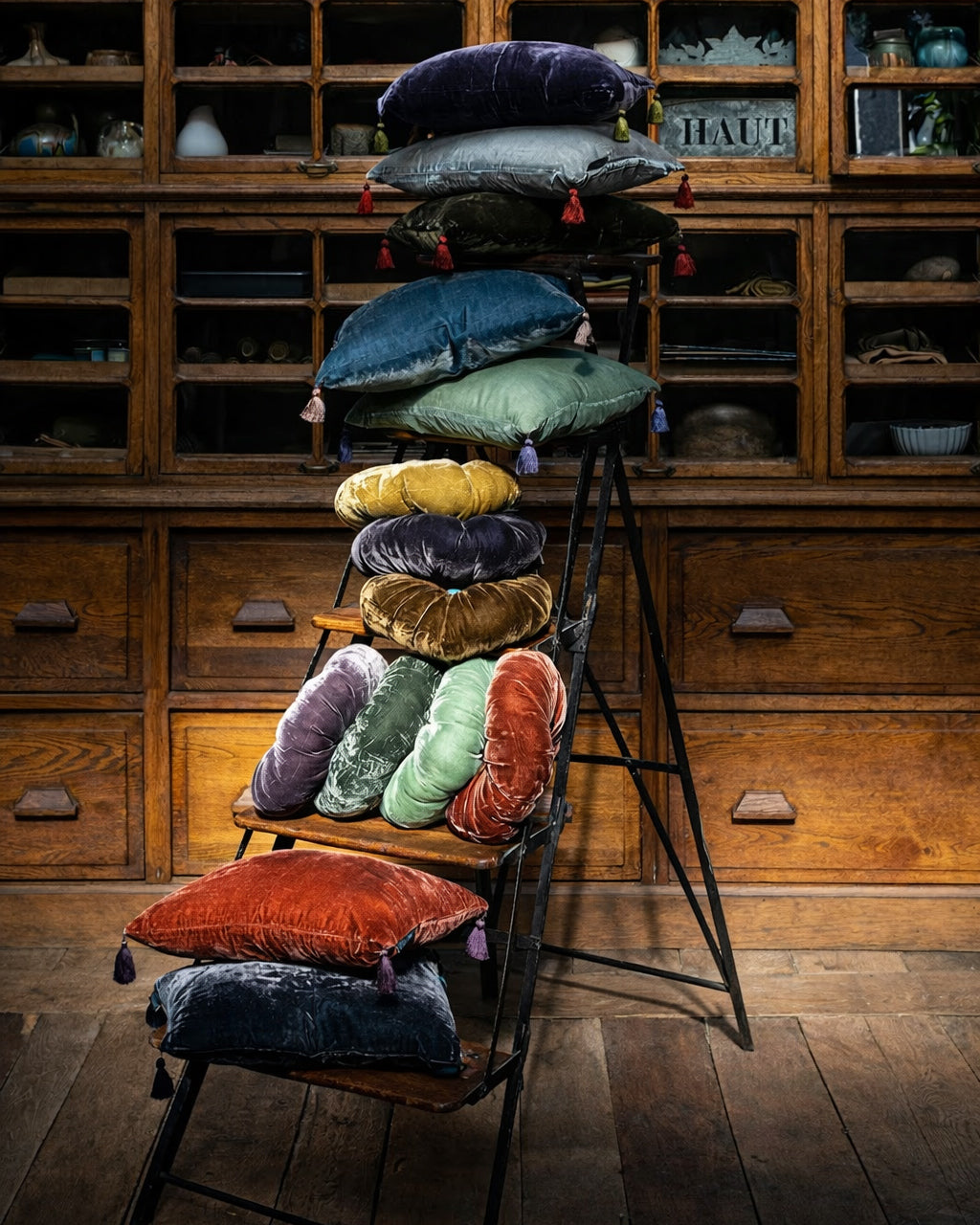 Colorful silk velvet velour pillows arranged on a ladder against a wooden cabinet background. Pillows by Monde Sauvage Paris.