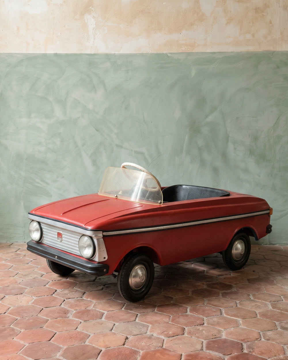 A vintage red Mokvich pedal car styled like a convertible roadster, featuring a clear windshield, black interior, and chrome details, parked on a terracotta tile floor against a textured green and beige plaster wall. Available via Wildschut Antiques.