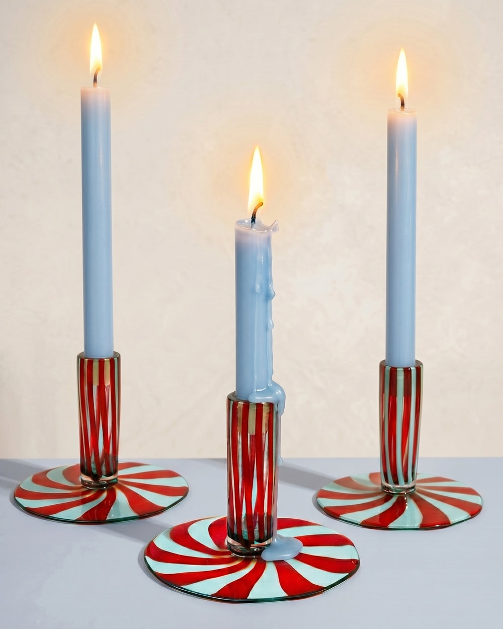 Three Murano glass candle holders with red and pale blue striped bases, each topped with a light blue burning candle, styled against a neutral background. Decorative Italian glassware available at Wildschut Antiques.