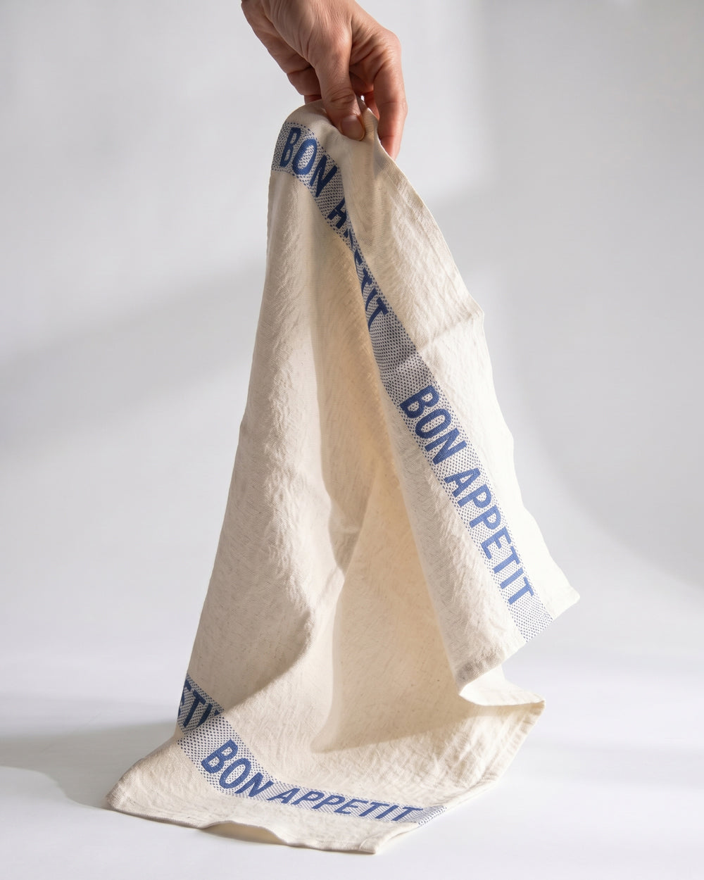 Hand holding a white linen napkin with blue text Bon Appetit. By Charvet Editions, available at Wildschut Antiques.