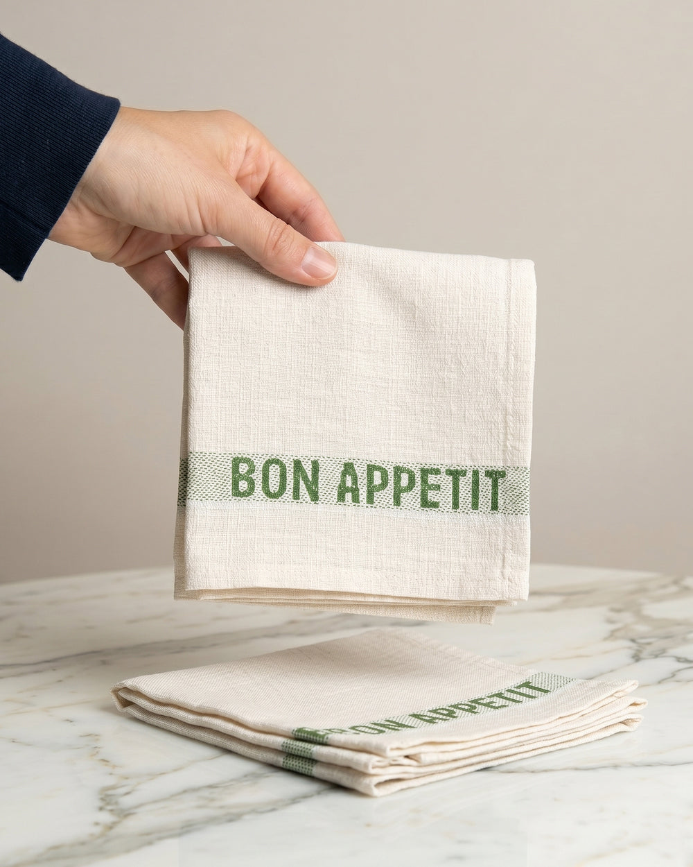 Hand holding a folded linen napkin with green 'Bon Appetit' text on a marble surface. By Charvet Editions, available at Wildschut Antiques.
