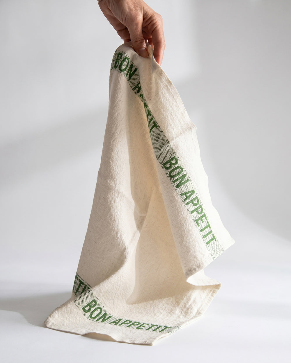 Hand holding a ecru white linen napkin with green text Bon Appetit. By Charvet Editions, available at Wildschut Antiques.