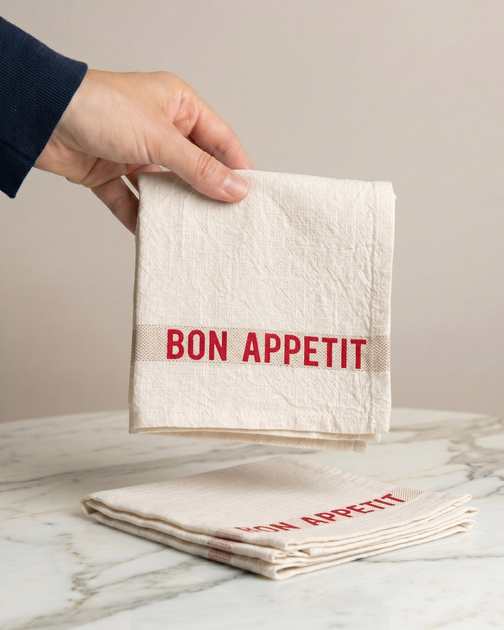 Hand holding a folded linen napkin with red 'Bon Appetit' text on a marble surface. By Charvet Editions, available at Wildschut Antiques.