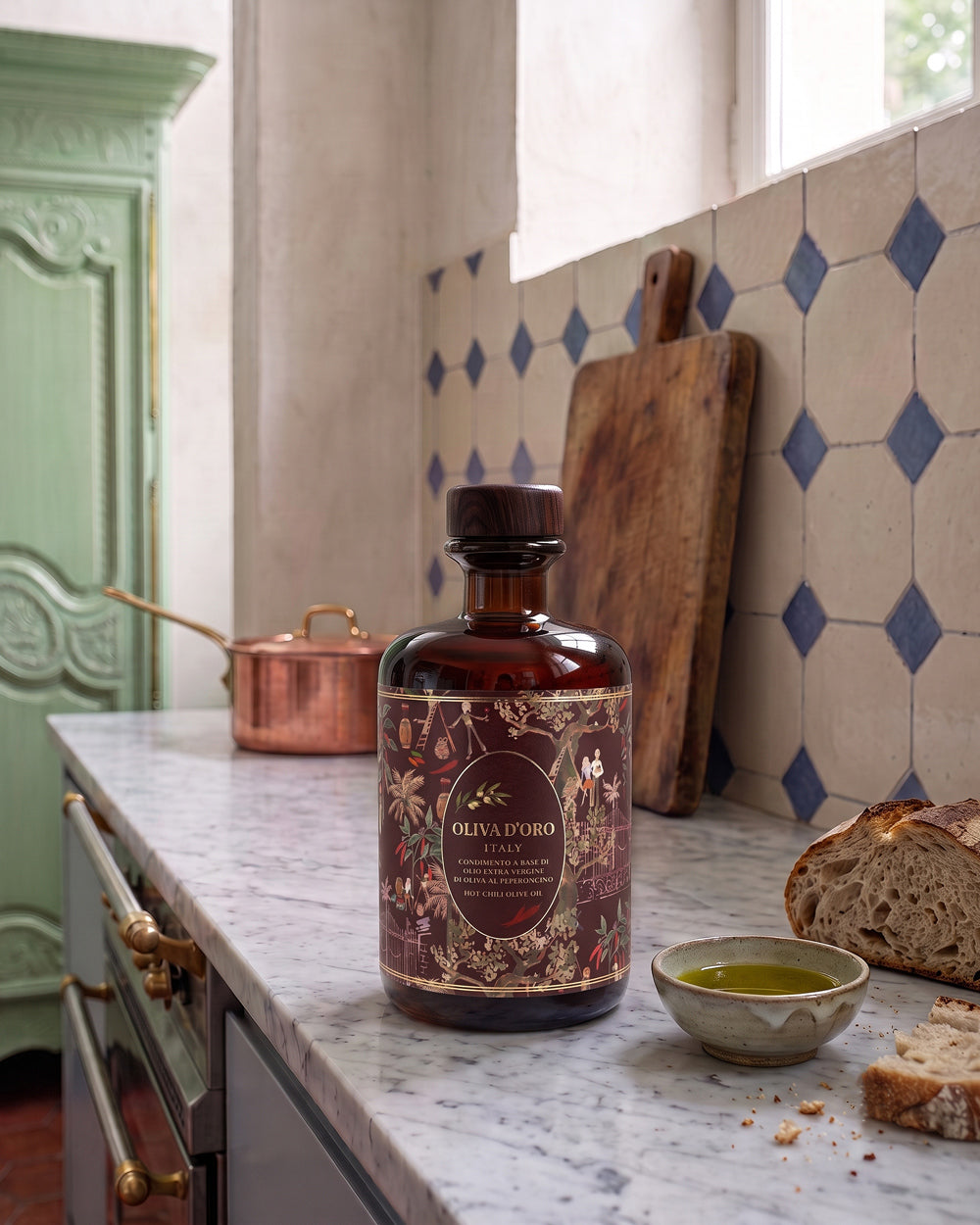 A bottle of olive oil from Oliva d'Oro stands on a marble kitchen counter, surrounded by rustic props including a cutting board, copper pan, bread, and a small bowl of oil. The background features a tiled wall with blue and beige diamond patterns.