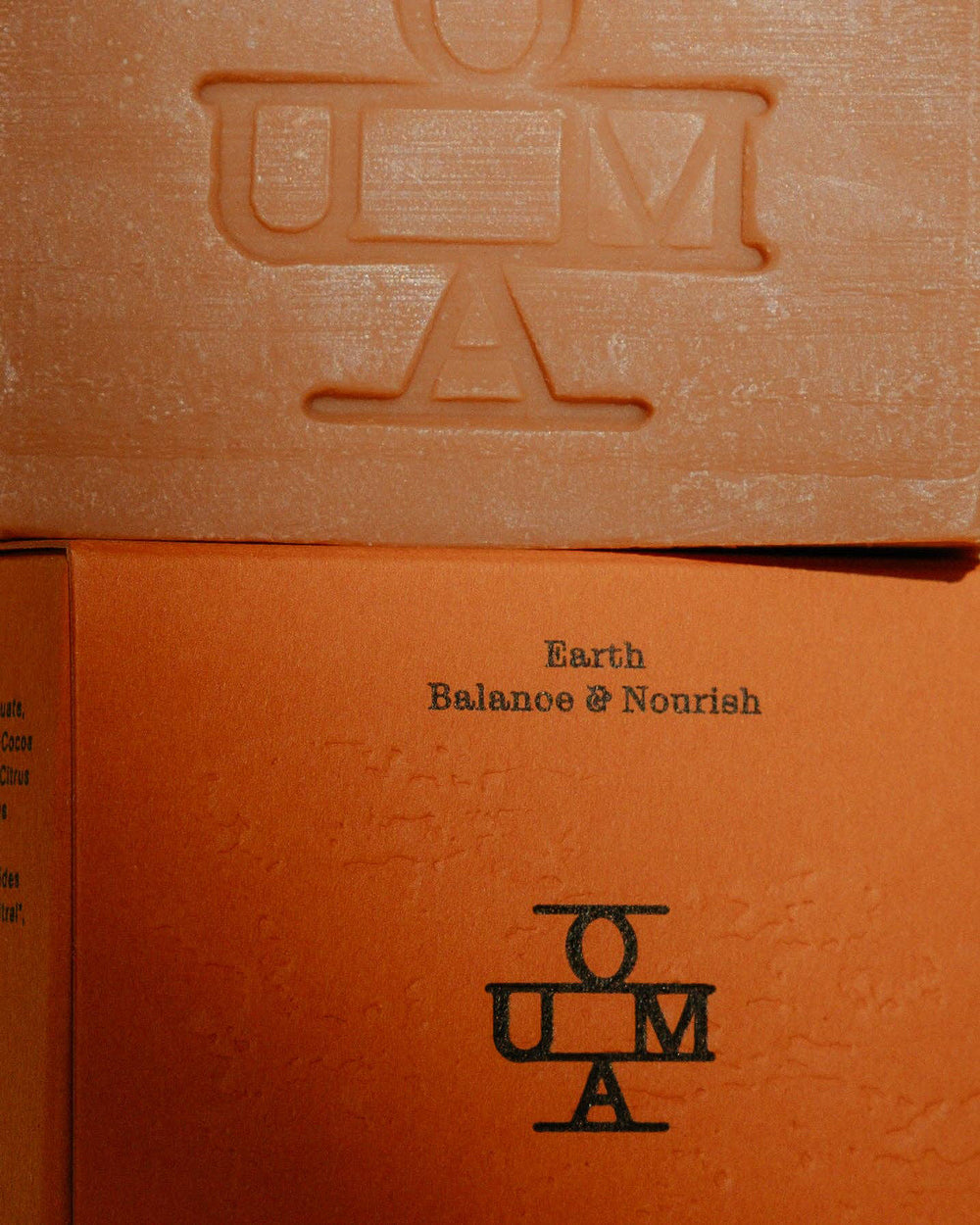 A close-up of a brown-red-colored soap bar with an embossed OUMA logo, positioned above its textured red packaging box labeled 'Earth Balance & Nourish'.