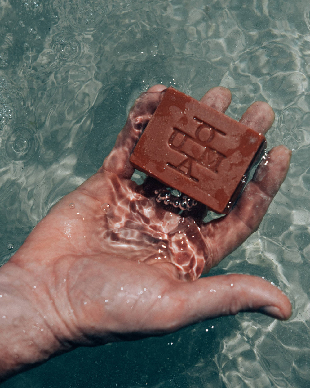 A hand in fresh water holding a bar of soap with an embossed logo that reads OUMA.