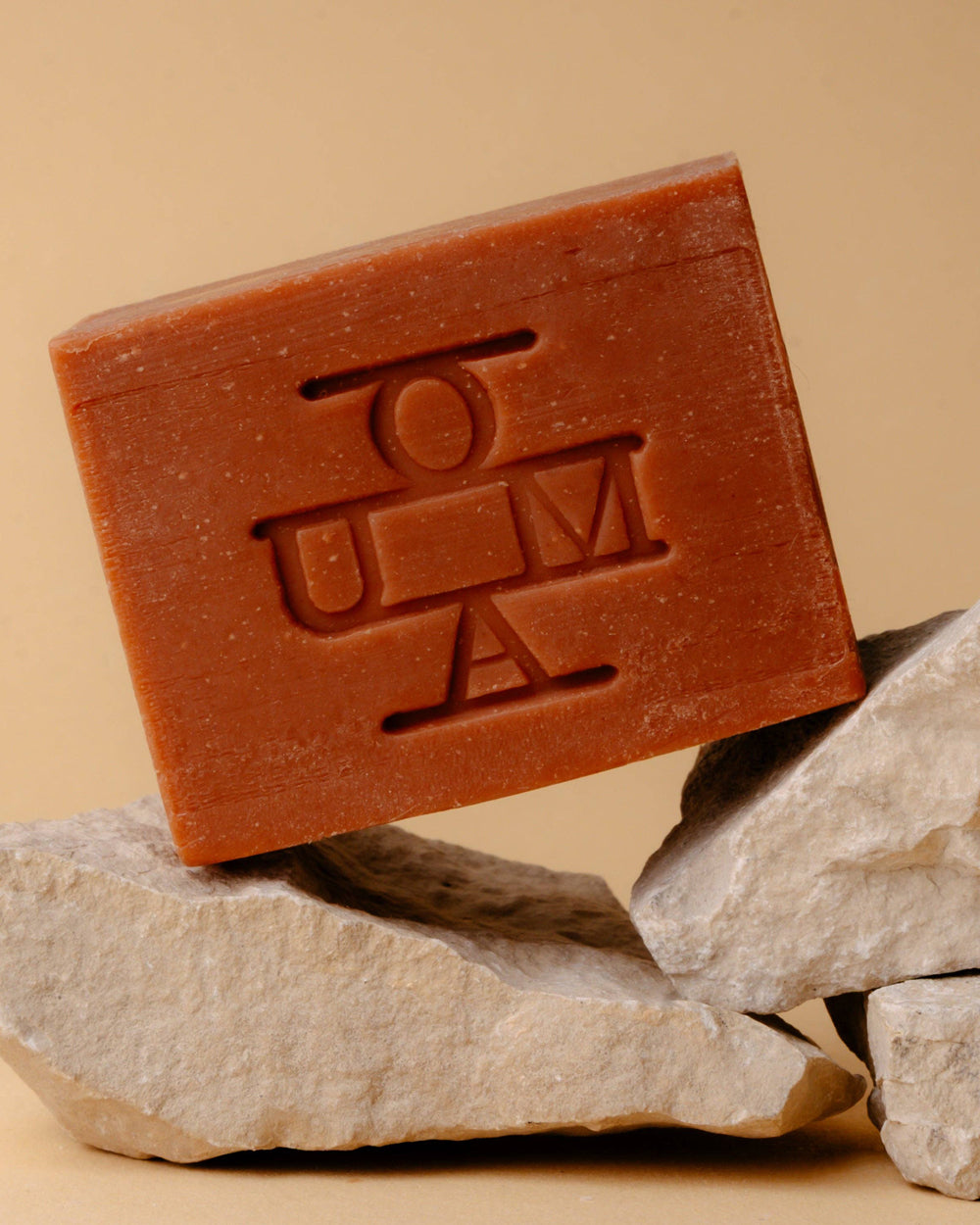 A terracotta-colored rectangular bar of soap with an embossed 'OUMA' logo, resting on stacked light gray stones against a solid beige background.