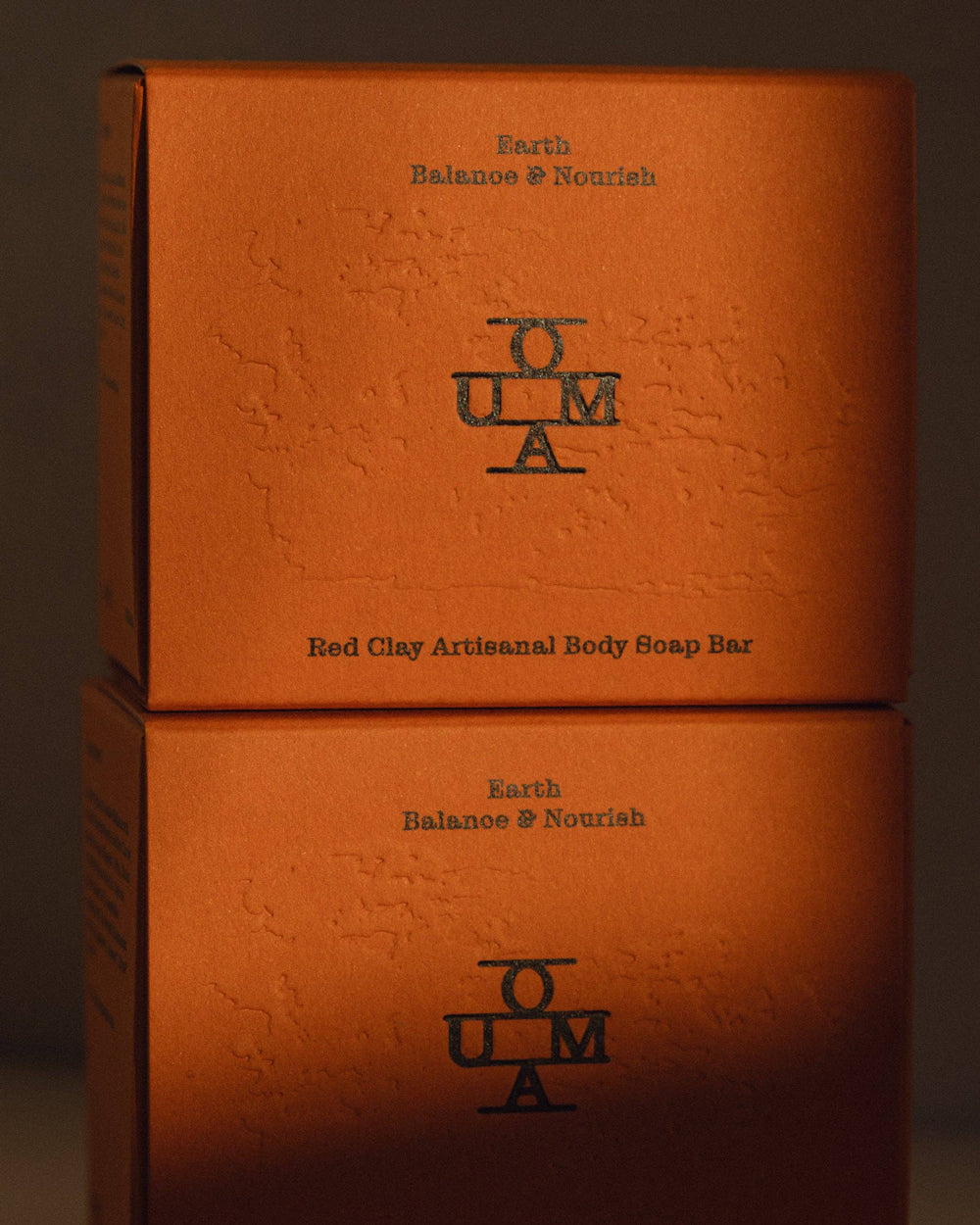 Two stacked orange-brown textured boxes of OUMA Earth Balance & Nourish Red Clay Artisanal Body Soap Bar, photographed in warm directional light against a dark background.